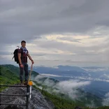 student on a hike standing on a rock with a hiking stick with clouds and expansive mountains behind him