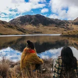 two student sitting on the bank of a glassy lake with mountains reflected in it and in the distance