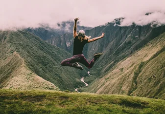 A woman jumps in the air with mountains and valleys in the foreground.