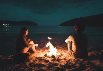 Two people sit by a camp fire on a beach at night.